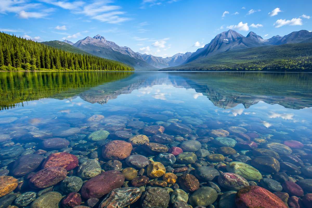 Crystal-clear mountain lake mcdonald reflecting forested peaks beneath a blue sky.